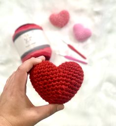 a hand holding a red crocheted heart next to yarn balls and knitting needles