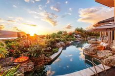 an outdoor swimming pool surrounded by rocks and plants with the sun setting in the background