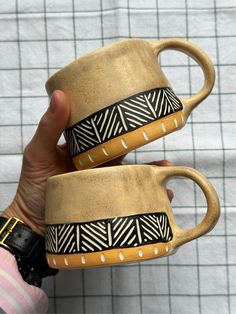 two handmade coffee mugs with black and white designs on them are held in front of each other