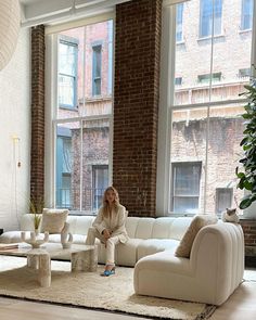 a woman sitting on a white couch in a living room with large windows and brick walls