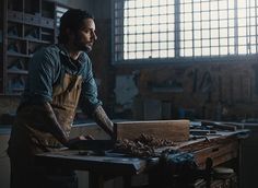 a man with tattoos working on wood in a room filled with windows and other tools