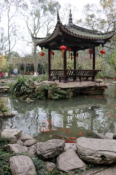 a gazebo in the middle of a pond surrounded by rocks and plants with red lanterns hanging from it