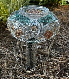 a glass candle holder sitting on the ground in front of some grass and bushes with flowers