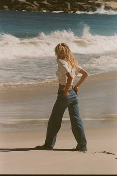 a woman standing on top of a sandy beach next to the ocean with her hands in her pockets