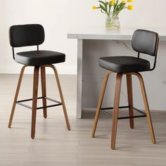 two black barstools with wooden legs in front of a white table and yellow flowers