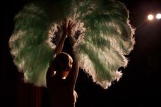 a woman is holding up a large green feathered fan in the dark with lights behind her