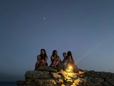 three women sitting on rocks near the ocean at night, one is holding a flashlight