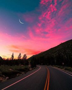 the sun is setting on an empty road with trees and mountains in the back ground