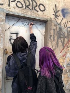 two people writing on a wall with purple hair and backpacks in front of them