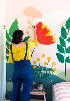 a woman in overalls painting a flower on a wall with flowers and leaves painted on it