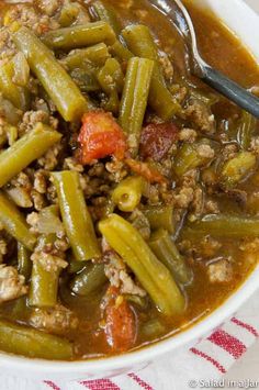 a white bowl filled with green beans, meat and tomato soup on top of a red and white checkered table cloth