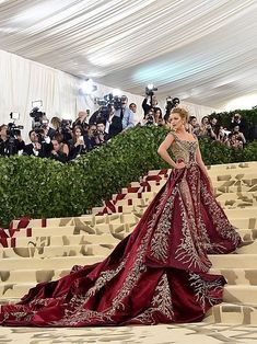 a woman in a red and gold gown standing on steps with cameras around her looking at the camera