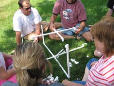 several people sitting on the grass playing with an object made out of sticks and pipes