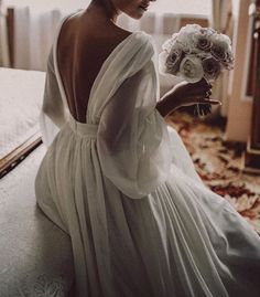 a woman in a white dress sitting on a bed holding a bridal bouquet and looking off to the side