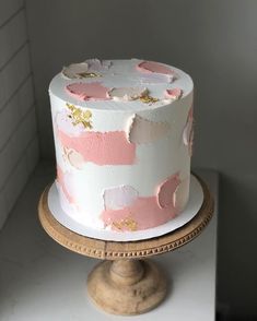 a white cake with pink and gold icing on a wooden stand next to a brick wall