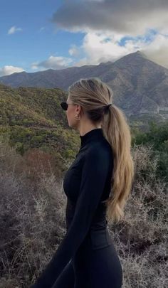 a woman with long hair and sunglasses is walking through the brush in front of mountains