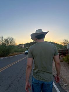 a man wearing a cowboy hat standing in the middle of an empty road at sunset