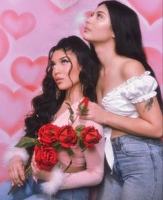 two beautiful women standing next to each other in front of a wall with hearts on it