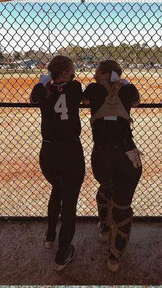 two women standing next to each other in front of a chain link fence and looking at a baseball field