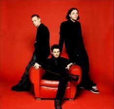 two men sitting on a red chair in front of a red wall and one man is wearing black