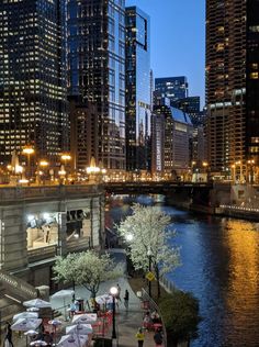 the city skyline is lit up at night with people walking on the sidewalk and tables in the foreground