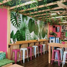 the inside of a restaurant with pink walls and green plants hanging from the ceiling, along with wooden tables and stools