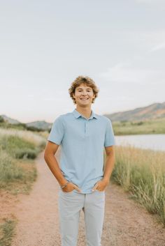 a young man standing on a dirt road in front of a body of water with his hands in his pockets