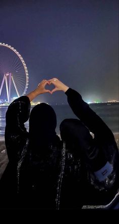 two people making a heart shape with their hands at night in front of the ferris wheel