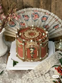 a red cake with gold decorations on it and a fan in the background next to some flowers