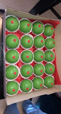 a box filled with green and red cupcakes on top of a table next to someone's feet