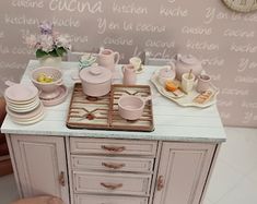 a dollhouse kitchen set with dishes and cups on the counter, next to a clock