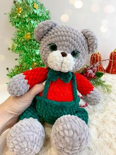 a crocheted teddy bear with green overalls and a red shirt sitting in front of a christmas tree