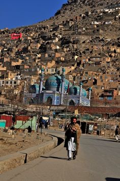 a man walking down a street in front of a large hill with buildings on it