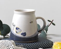 a white and blue coffee mug sitting on top of a table next to yellow flowers