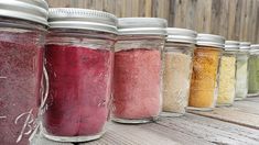 seven jars filled with different colored powders on a wooden table next to a fence