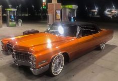 an orange classic car parked in front of a gas station at night with its lights on