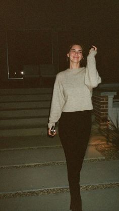 a woman in black pants and a white sweater is walking down the street at night