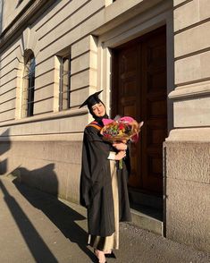 a woman in a graduation gown holding flowers and a teddy bear on the side of a building