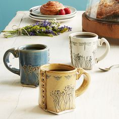 three coffee mugs sitting on top of a table next to some pastries and fruit