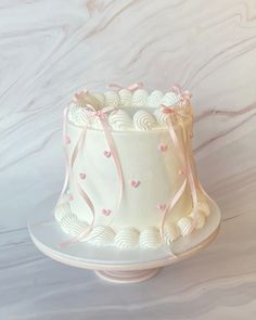 a white cake with pink ribbon and hearts on it sitting on a stand in front of a marble wall