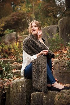 a woman is sitting on some rocks and talking on her cell phone while wearing a shawl