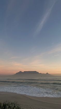 the sun is setting over an ocean with mountains in the distance and waves crashing on the beach