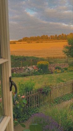 an open window looking out onto a field with flowers and trees in the distance on a cloudy day