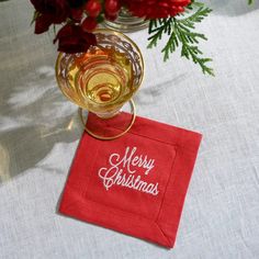 a red napkin with merry christmas written on it next to a wine glass and flowers