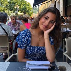 a woman sitting at an outdoor table with her hand on her chin and looking into the camera