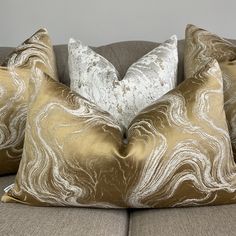 three pillows sitting on top of a couch with gold and white designs in the middle