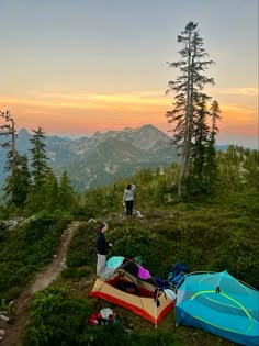 two people standing on top of a mountain next to tents and backpacks at sunset