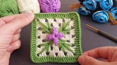 a hand holding a crochet square with flowers on it and yarn in the background