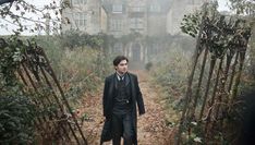 a man in a long black coat is walking down a dirt path with an old house in the background