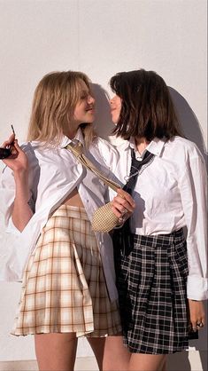 two young women standing next to each other wearing skirts and ties, one holding a cell phone